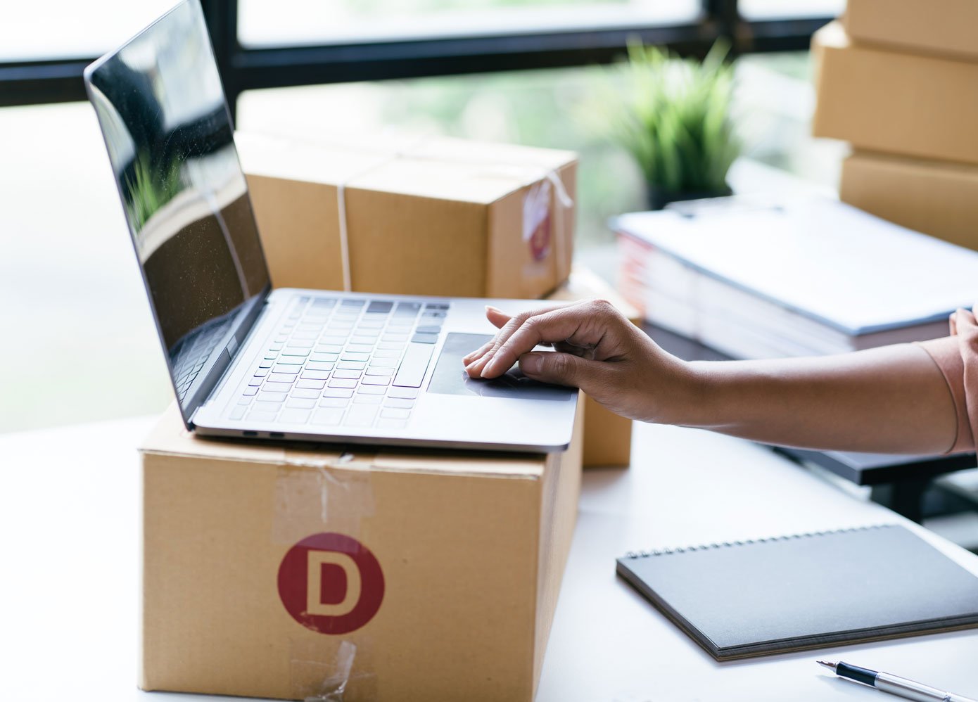 Person managing a Shopify dropshipping business using a laptop, with shipping boxes and inventory in the background.