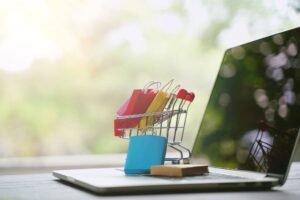 A miniature shopping cart filled with colourful shopping bags sitting on a laptop, symbolizing online shopping and e-commerce growth in 2025.